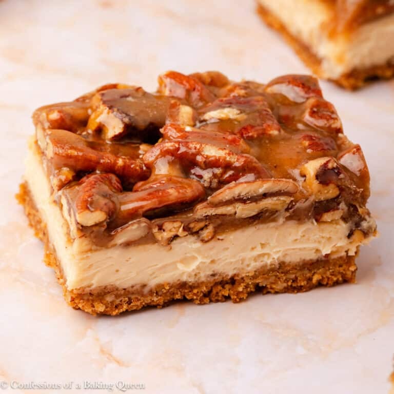 A close-up of pecan pie cheesecake bars with a graham cracker crust, creamy cheesecake layer, and caramelized pecans on top, set on a light marble surface.
