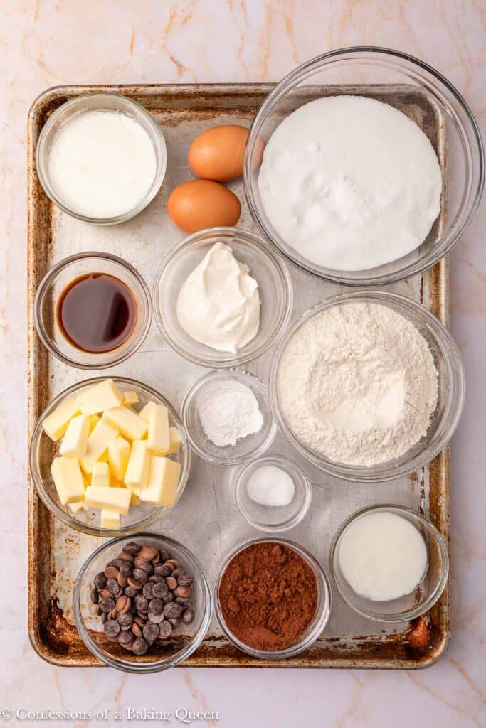 A baking tray holds bowls of ingredients including two eggs, sugar, flour, cocoa powder, butter cubes, chocolate chips, sour cream, vanilla extract, baking soda, salt, milk, and another liquid.
