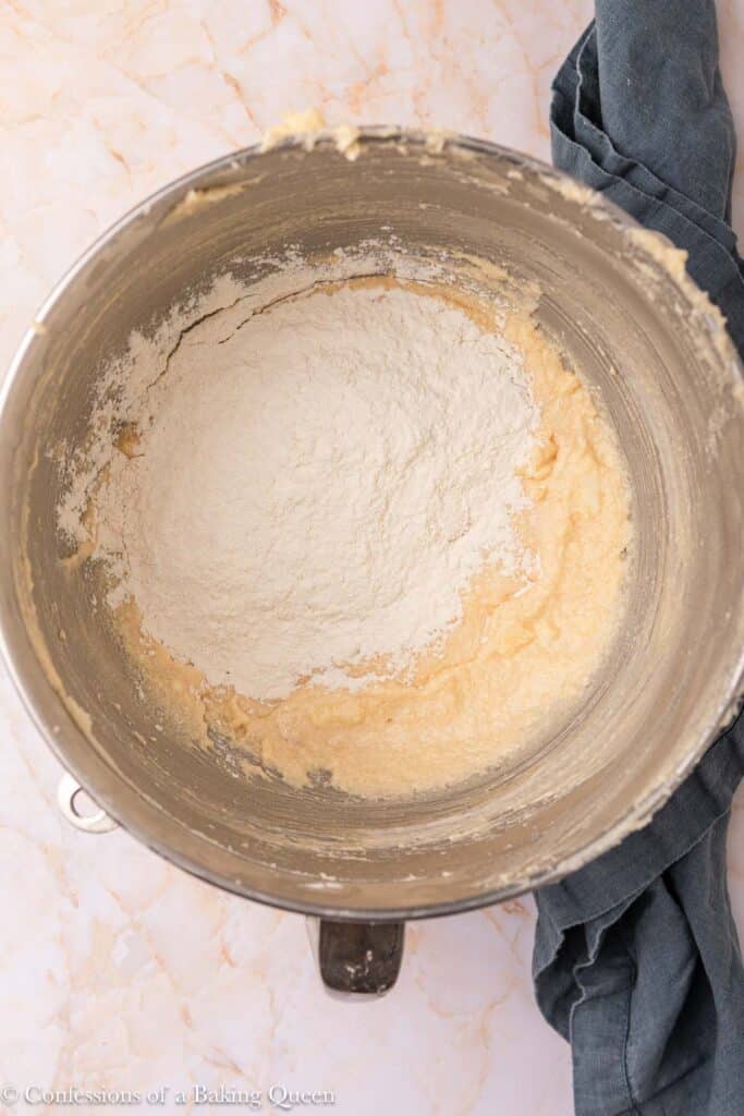 A metal mixing bowl containing partially mixed batter with a mound of flour on top, resting on a light marble countertop next to a dark kitchen towel.
