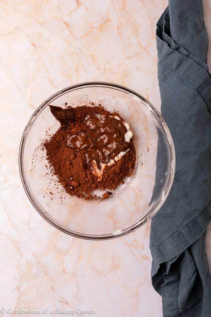 A glass mixing bowl with cocoa powder, melted chocolate, and cream cheese on a marble surface. A dark gray kitchen towel lies beside the bowl.