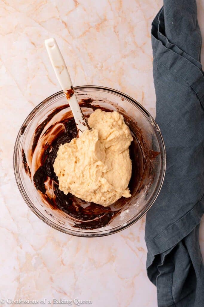 A glass bowl with chocolate and light-colored batter being mixed together with a white spatula, placed on a marble surface next to a folded blue-gray cloth.
