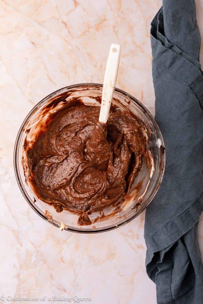 A glass bowl filled with chocolate brownie batter and a white spatula rests on a marble surface next to a crumpled dark gray cloth.