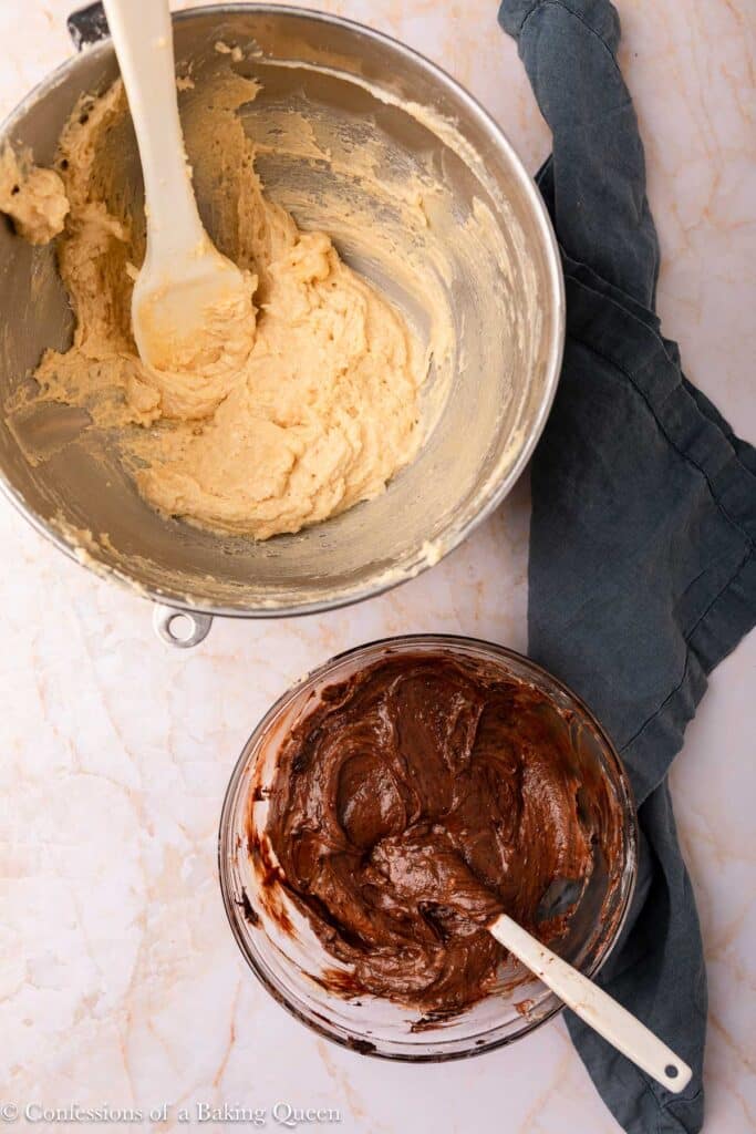Two mixing bowls on a marble surface: the top metal bowl contains a light-colored batter with a spatula, and the bottom glass bowl holds chocolate batter with a spatula. A dark kitchen towel is beside the bowls.