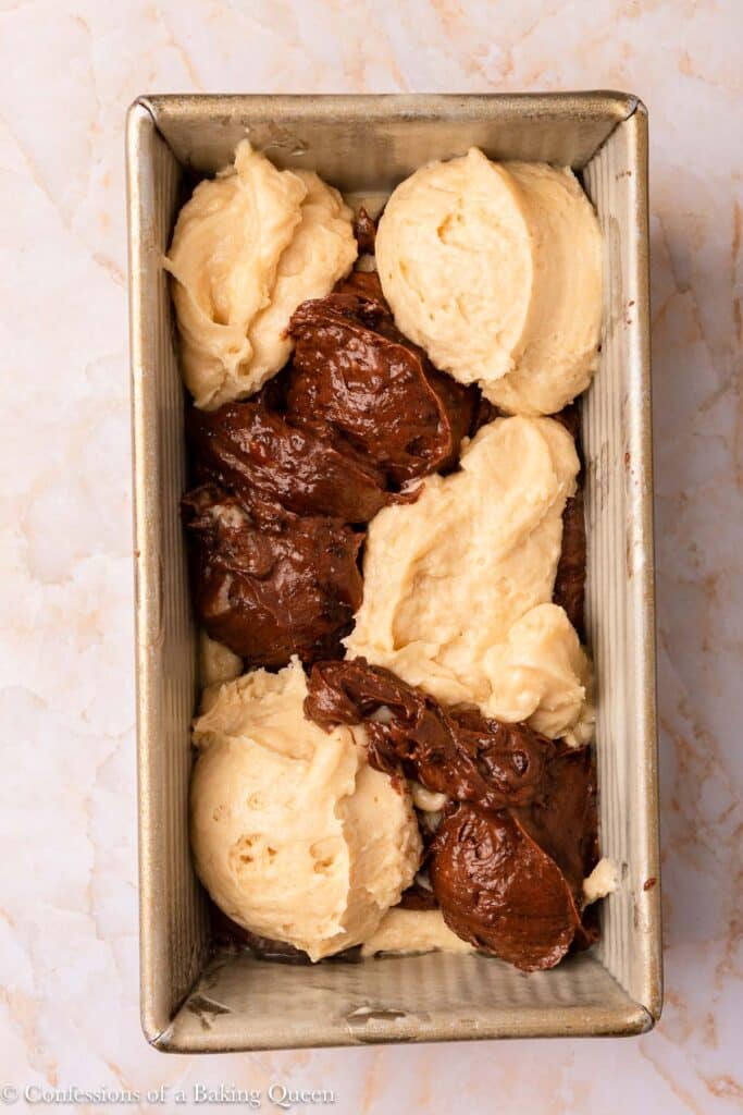 A rectangular metal baking pan filled with alternating dollops of light vanilla and dark chocolate cake batter, ready to be swirled or baked, on a light marble surface.