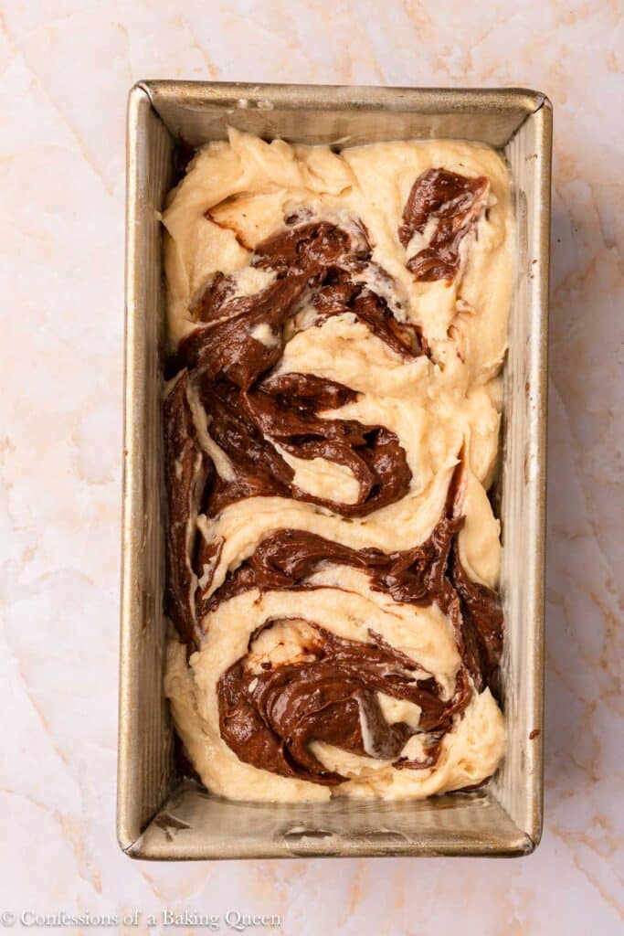 A rectangular metal loaf pan filled with unbaked marble loaf cake batter, swirled with chocolate, sits on a light marble surface.