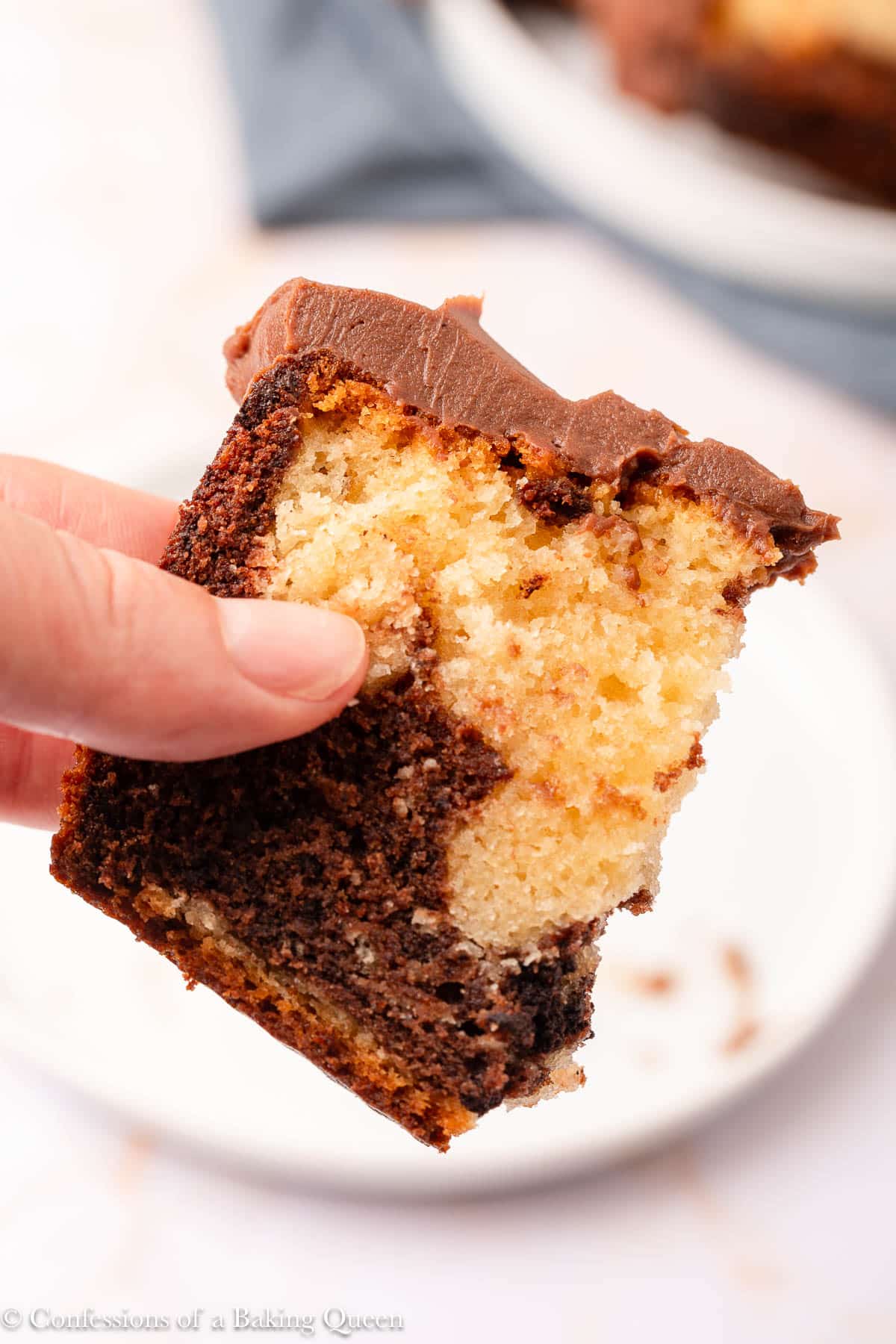 A hand holding a slice of marble cake with swirls of chocolate and vanilla, topped with a thick layer of chocolate frosting. A white plate with crumbs is visible in the background.