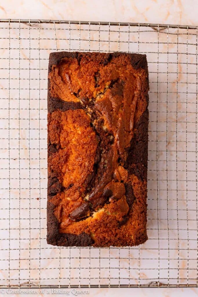 A loaf of marble cake with swirls of chocolate and vanilla, resting on a metal cooling rack over a light marble surface.