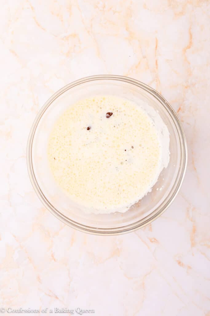 A glass bowl filled with a creamy, pale yellow liquid with a few small dark specks, sitting on a light marble countertop.
