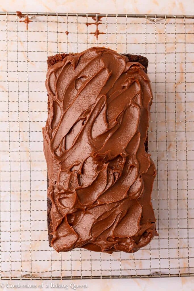 A loaf of chocolate cake with thick, swirled chocolate frosting sits on a wire cooling rack over a light-colored surface.