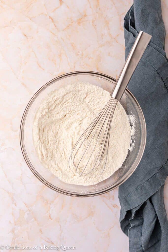 A glass bowl filled with flour and a metal whisk on a light marble surface, with a dark gray kitchen towel nearby.