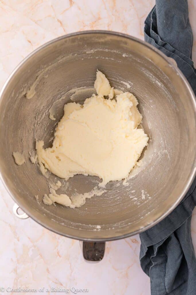 A metal mixing bowl with creamed butter and sugar sits on a marble surface next to a folded dark gray kitchen towel.