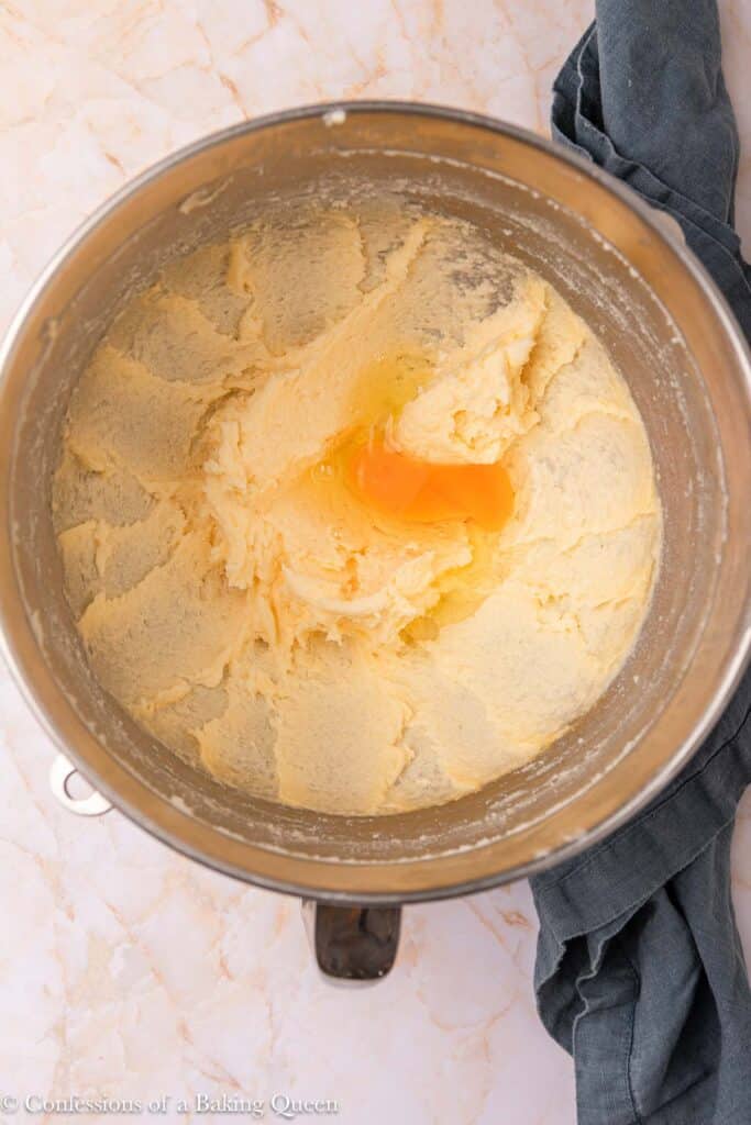 A metal mixing bowl contains creamed butter and sugar with a raw egg cracked in the center, sitting on a marble countertop next to a dark cloth.