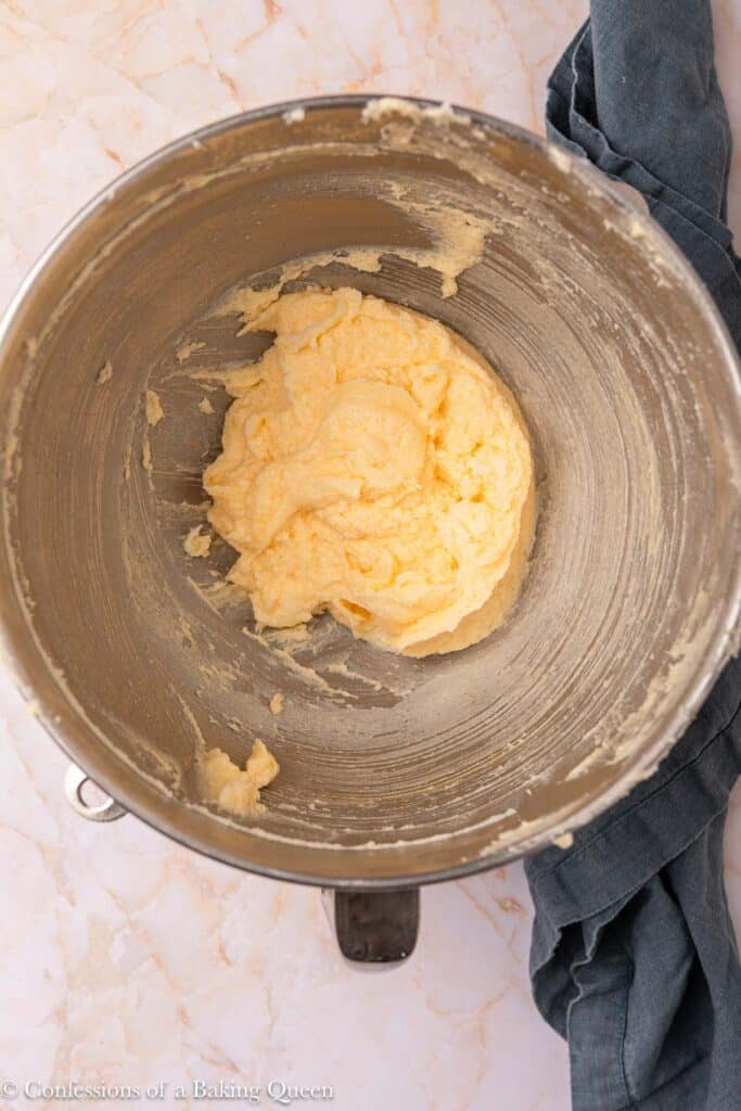 A metal mixing bowl containing creamed butter and sugar sits on a light marble surface with a dark cloth partially visible to the side.