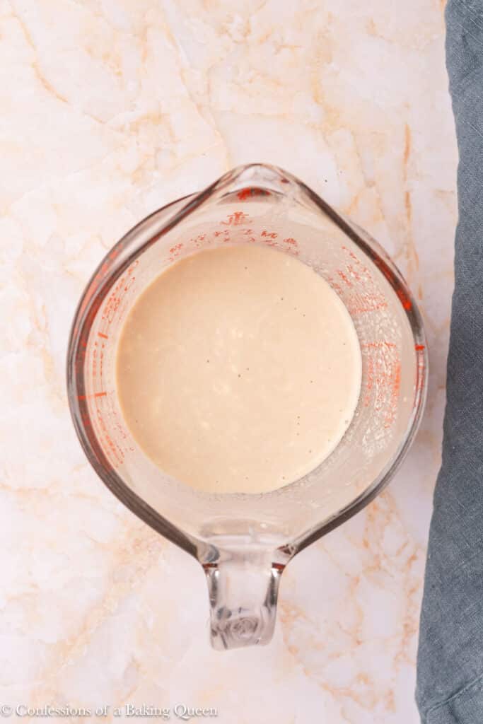 A glass measuring cup filled with a creamy beige liquid sits on a light marble surface next to a gray cloth.