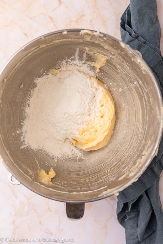A metal mixing bowl containing creamy batter and flour sits on a marble surface with a dark cloth partially visible to the right.