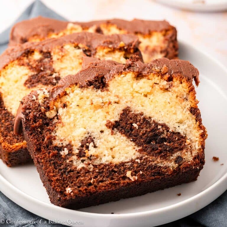 Two slices of marble loaf cake with chocolate and vanilla swirls, topped with a layer of chocolate frosting, served on a white plate.