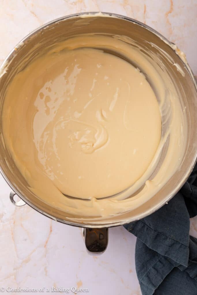 A metal mixing bowl filled with smooth, creamy cheesecake batter sits on a light marble surface, next to a dark cloth.