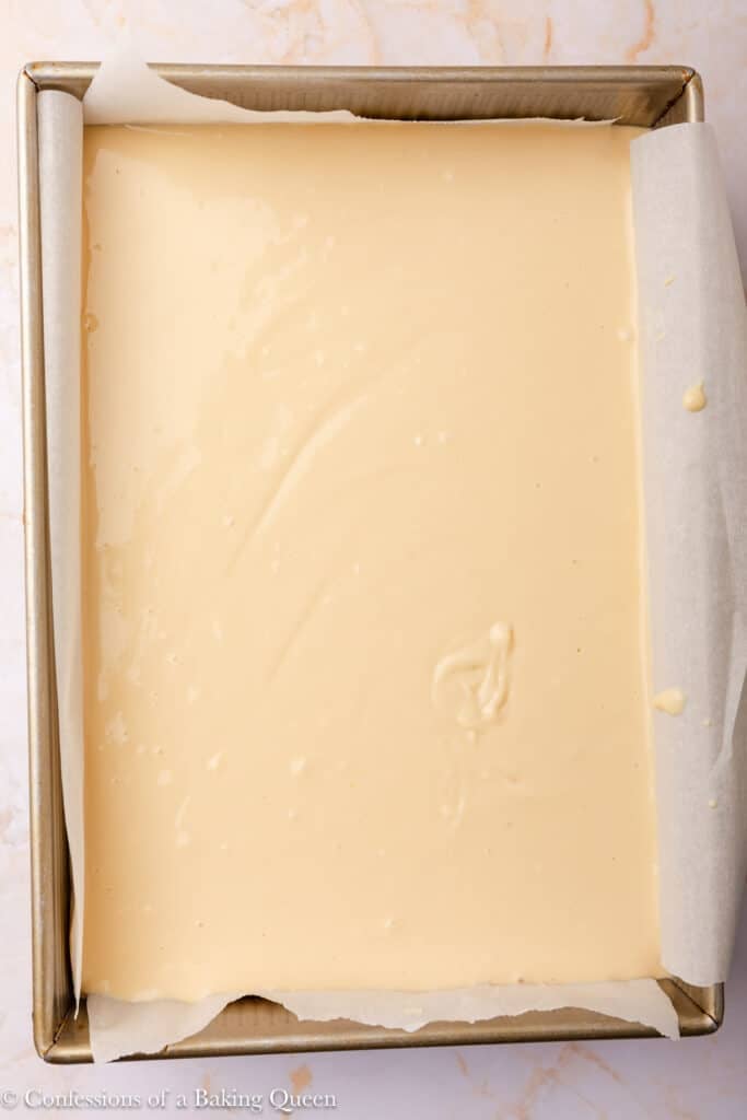 A rectangular metal baking pan lined with parchment paper is filled with smooth, pale yellow batter, ready to be baked. The pan sits on a light-colored marble surface.