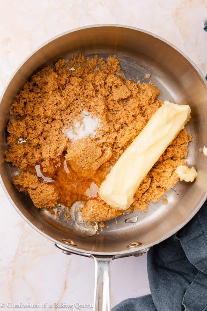 A saucepan contains a stick of butter, brown sugar, salt, and a bit of vanilla extract, all partially mixed together, sitting on a light-colored countertop next to a dark cloth.