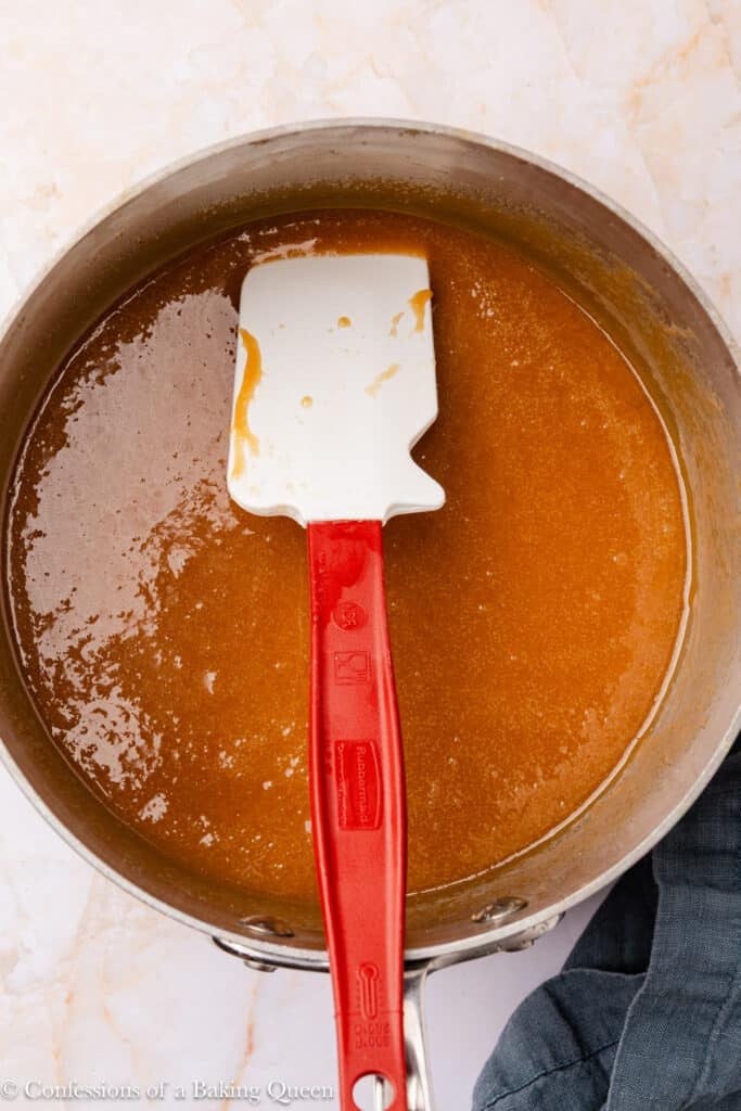 A saucepan filled with caramel sauce being stirred by a white and red silicone spatula, resting on a light marble surface.