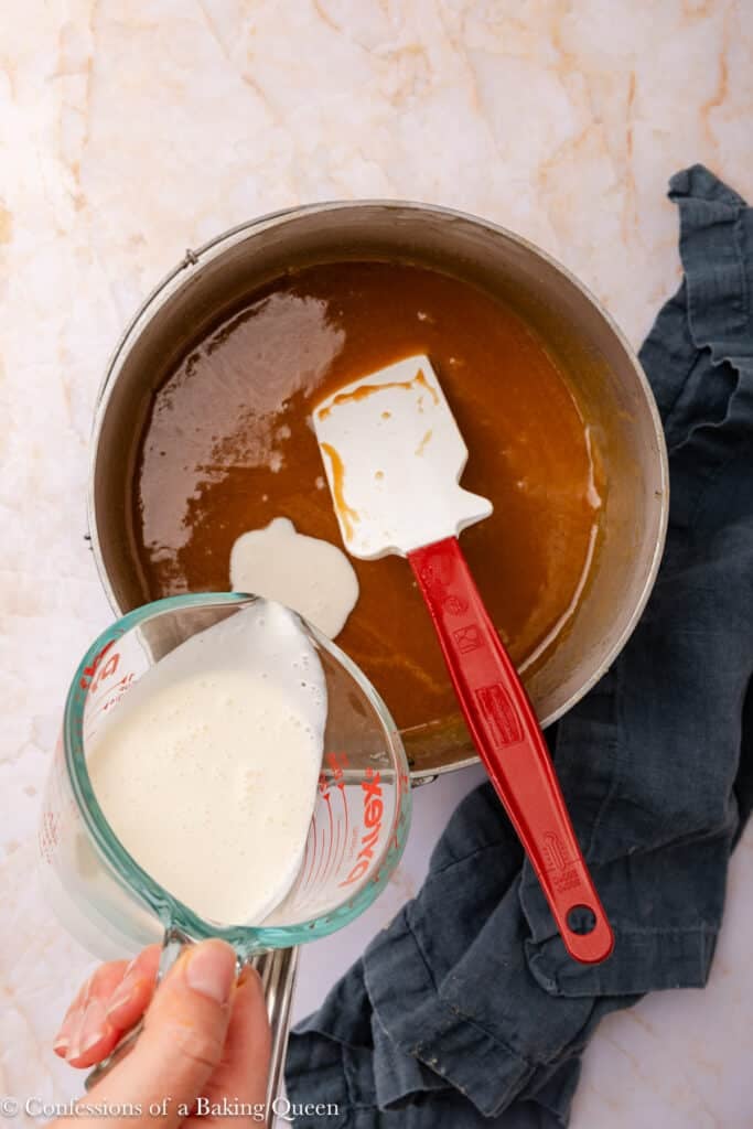 A hand pours cream from a glass measuring cup into a saucepan filled with caramel sauce, using a spatula to stir. A dark cloth is placed beside the saucepan on a light marble surface.