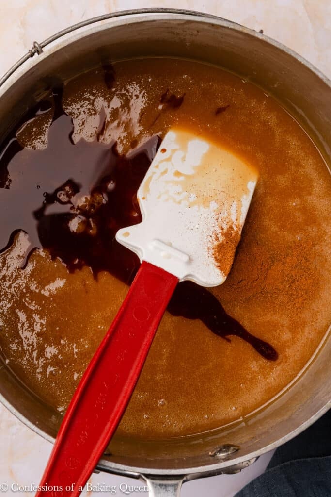 A pot filled with a light brown liquid mixture being stirred with a white spatula with a red handle; dark vanilla extract and cinnamon are visible on the surface.
