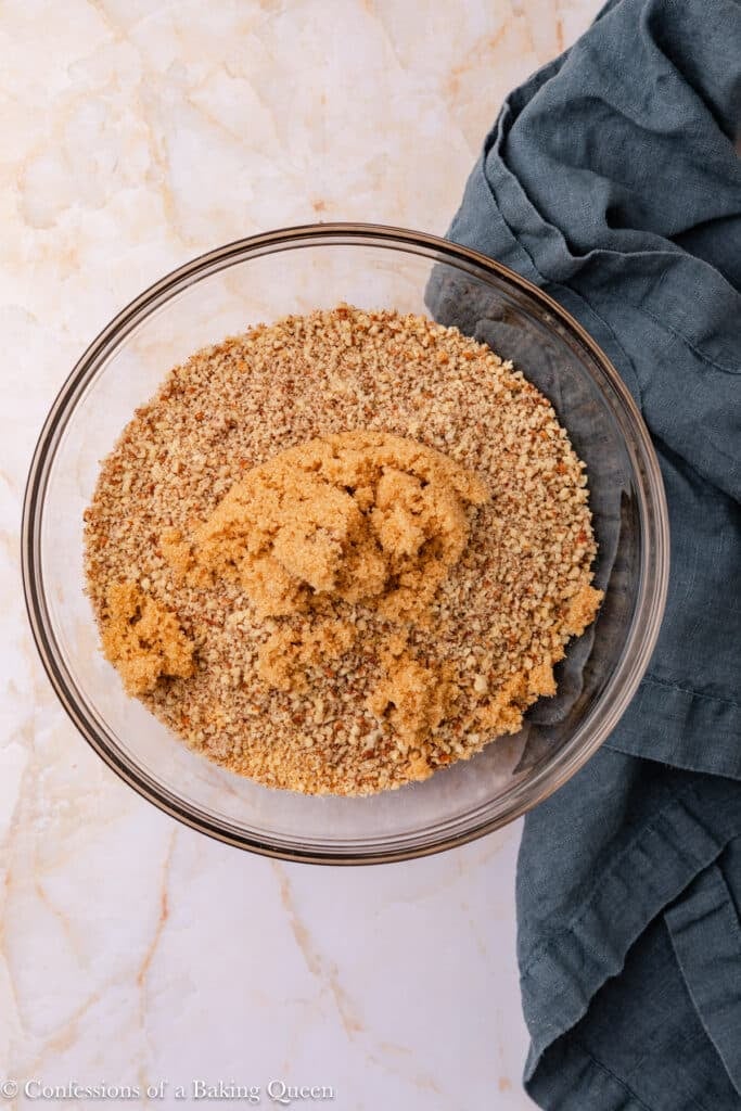 A glass bowl filled with finely ground nuts and brown sugar sits on a marble surface next to a folded dark gray cloth.