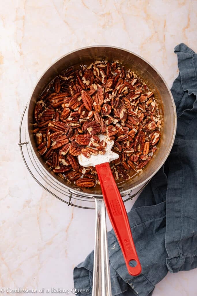A saucepan filled with pecans sits on a wire rack, with a red-handled spatula resting inside. A blue kitchen towel lies next to the saucepan on a light marble surface.