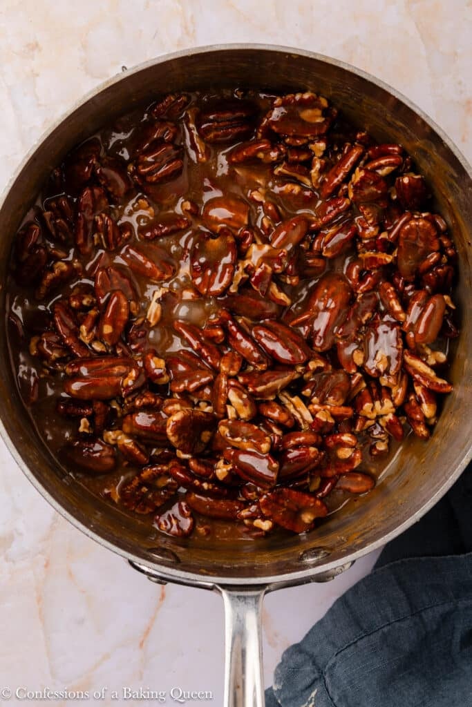 A saucepan filled with glossy, caramel-coated pecans sits on a light marble surface, with part of a dark cloth visible under the pan's handle.