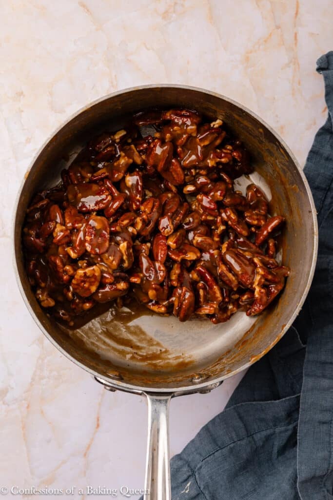 A saucepan filled with candied pecans coated in a glossy brown caramel sauce, resting on a light marble surface with a dark cloth nearby.