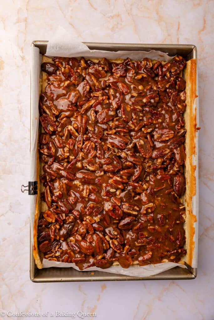 A rectangular baking pan lined with parchment paper holds a golden brown pecan pie bar with a glossy caramelized pecan topping, resting on a light marble surface.