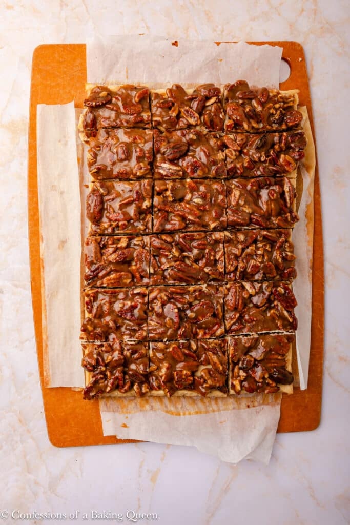 A batch of pecan bars cut into squares sits on parchment paper atop a brown cutting board, showing a caramelized nut topping over a light crust.