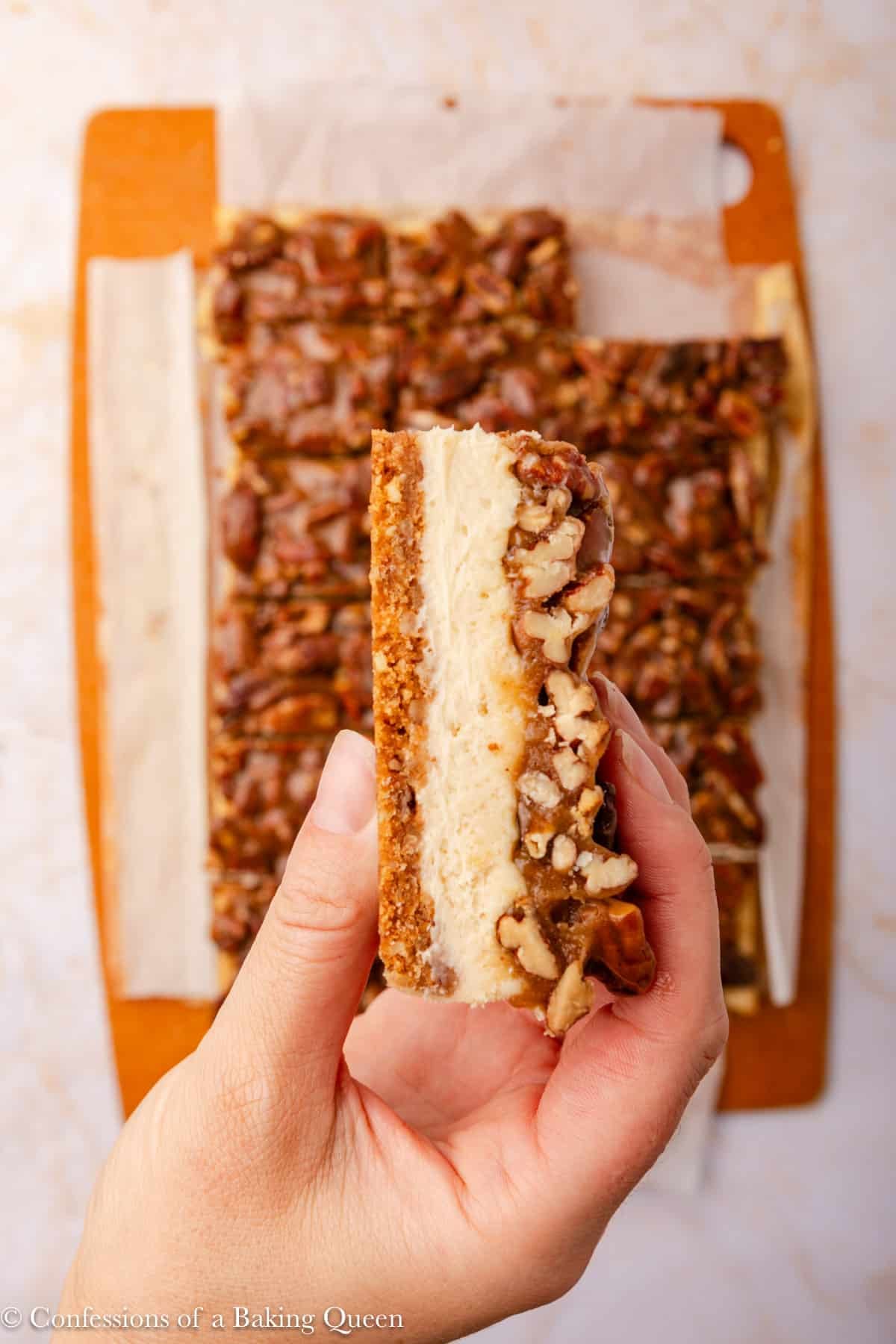 A hand holds up a layered dessert bar with a crumbly crust, creamy cheesecake middle, and a pecan topping. More bars are visible on parchment paper in the background.