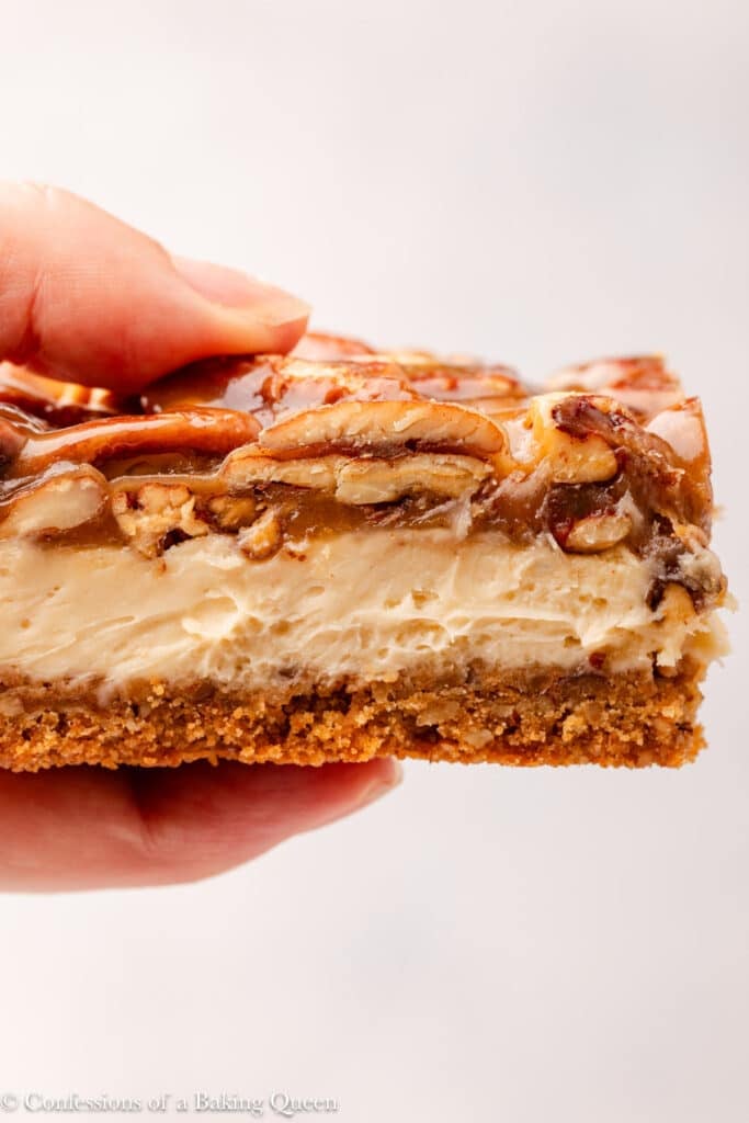 A close-up of a hand holding a pecan pie cheesecake bar, showing layers of graham cracker crust, creamy cheesecake, and glossy pecan topping.