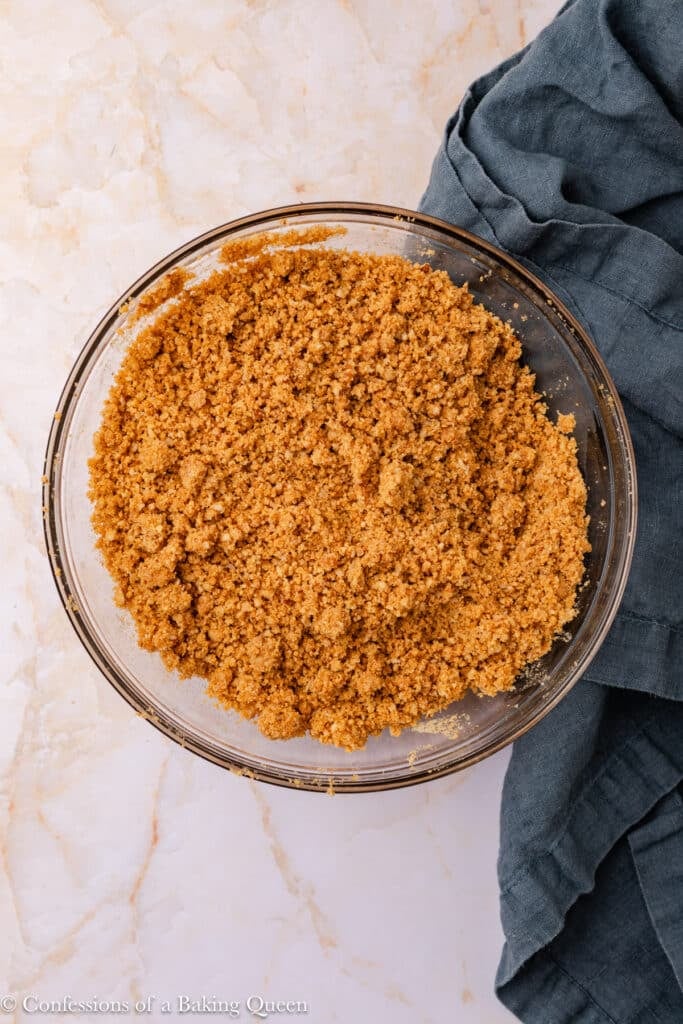 A glass bowl filled with crumbly graham cracker crust mixture sits on a light marble surface next to a dark gray fabric napkin.