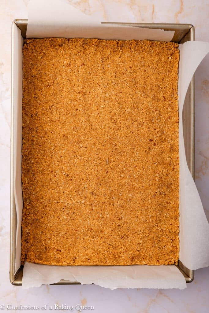 A rectangular metal baking pan lined with parchment paper holds an even layer of golden-brown crumb crust, pressed flat and ready for baking.