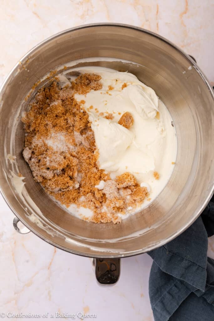 A mixing bowl containing cream cheese, brown sugar, and granulated sugar, ready to be blended together. The bowl sits on a marble countertop with part of a dark cloth nearby.