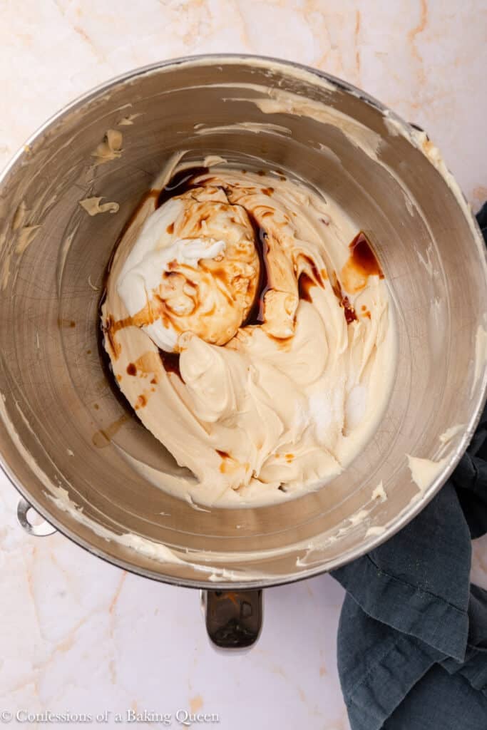 A metal mixing bowl containing creamy batter with swirls of vanilla extract and sugar on top, resting on a marble countertop with a dark cloth nearby.