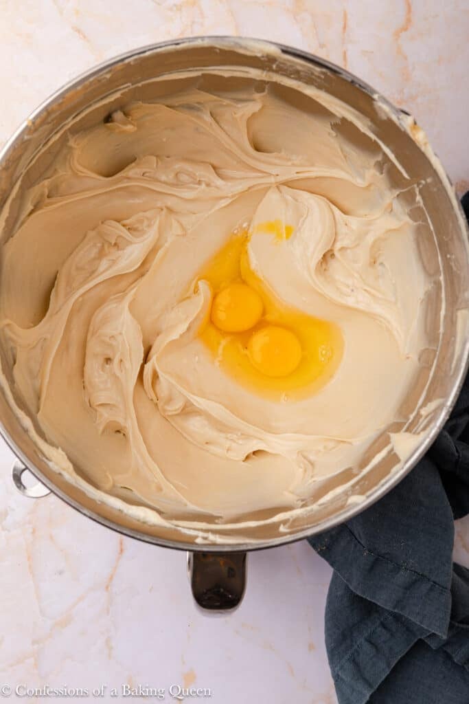 A metal mixing bowl filled with creamy batter and two raw eggs on top, ready to be mixed. The bowl is placed on a marble countertop with a dark cloth nearby.