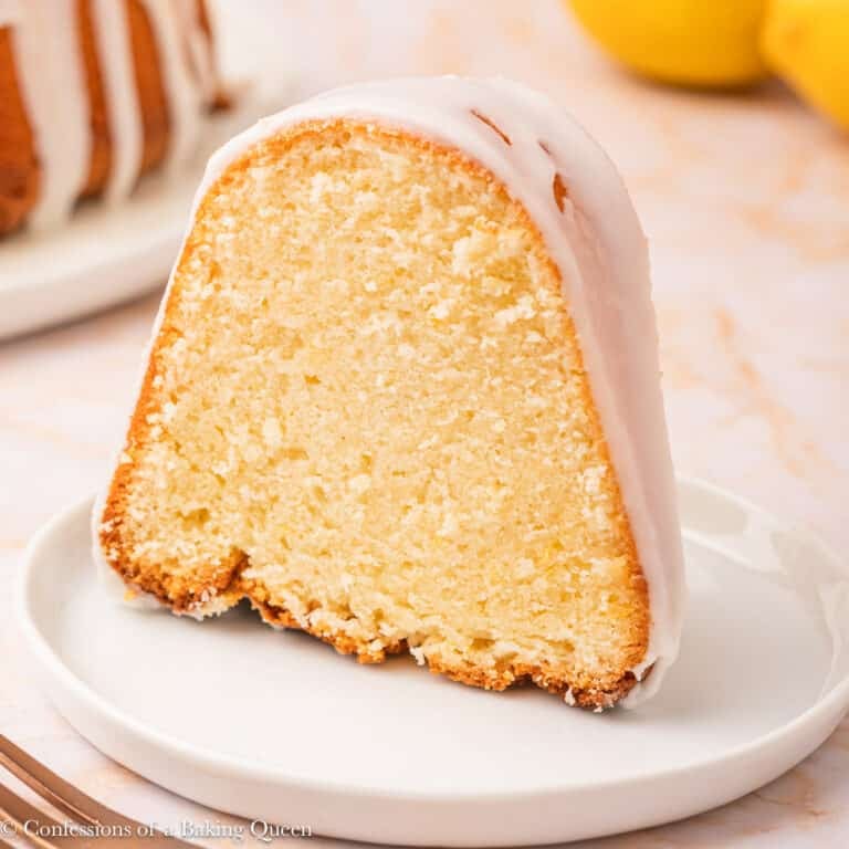 A close-up of a slice of lemon cream cheese bundt cake with a light glaze on top, served on a white plate. The texture of the cake looks moist and dense, and part of the whole cake is visible in the background.
