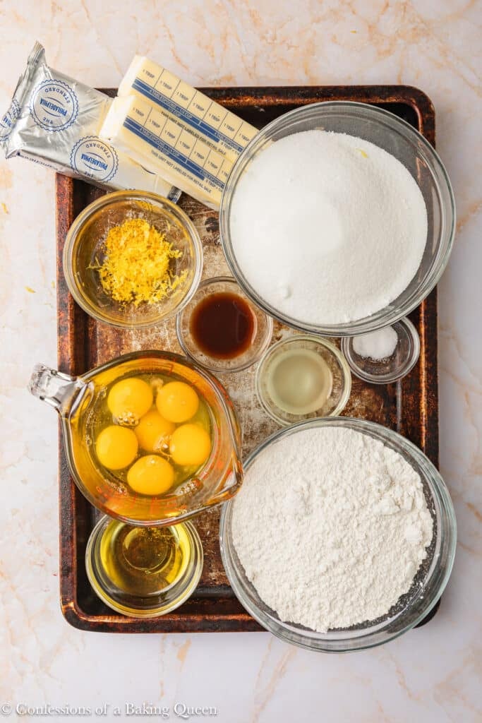 A baking tray holds bowls of flour, sugar, lemon zest, eggs, oil, vanilla, lemon juice, salt, and two sticks of butter, arranged neatly on a marble surface.