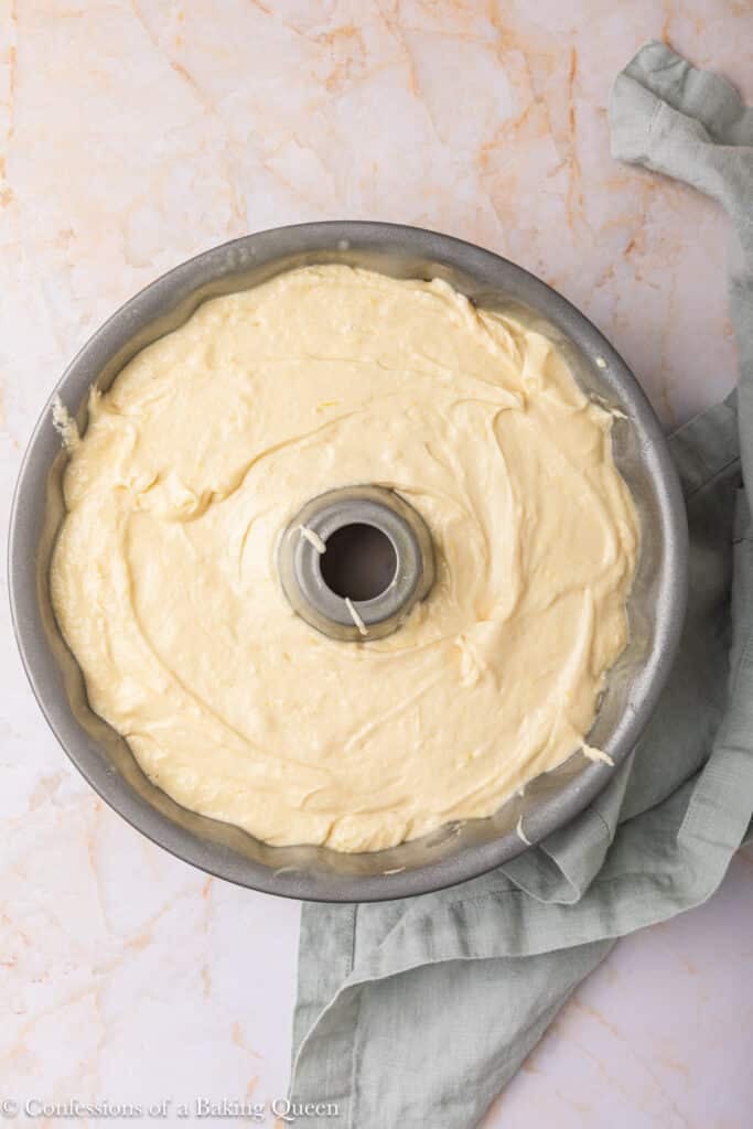 A round bundt pan filled with smooth, unbaked cake batter sits on a marble surface with a light green kitchen towel partially under the pan.