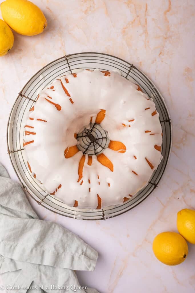 A lemon cream cheese pound cake sits on a round wire rack atop a marble surface, surrounded by whole lemons and a light gray cloth napkin. The bundt cake is topped with a thick layer of white icing.