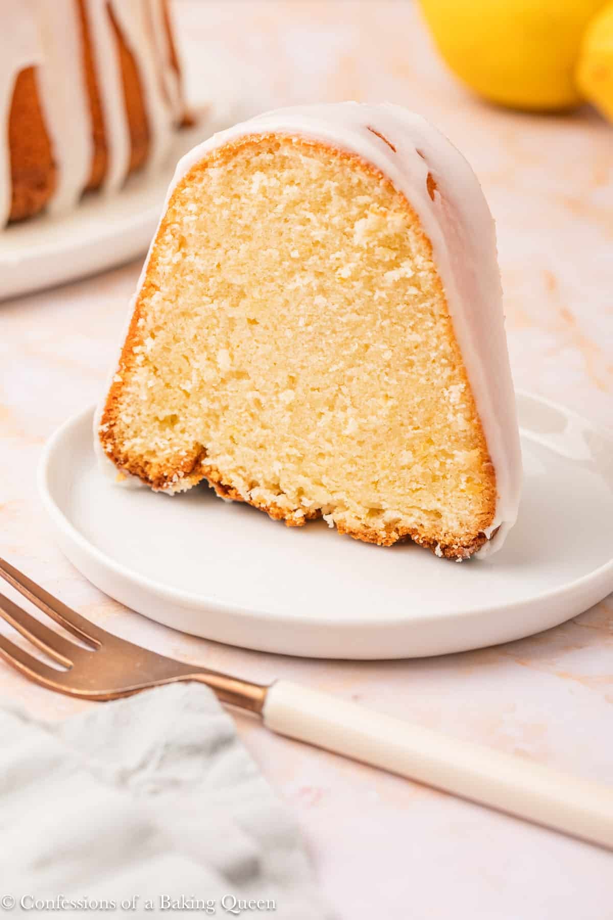 A single slice of bundt cake with white icing sits on a small white plate next to a fork. The cake has a moist, dense texture and a golden brown edge. The background shows part of the whole bundt cake and some lemons.