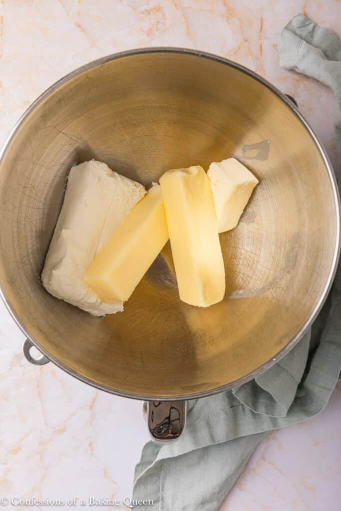 A metal mixing bowl contains two sticks of butter and a block of cream cheese on a light marble countertop, with a green cloth partially visible on the side.