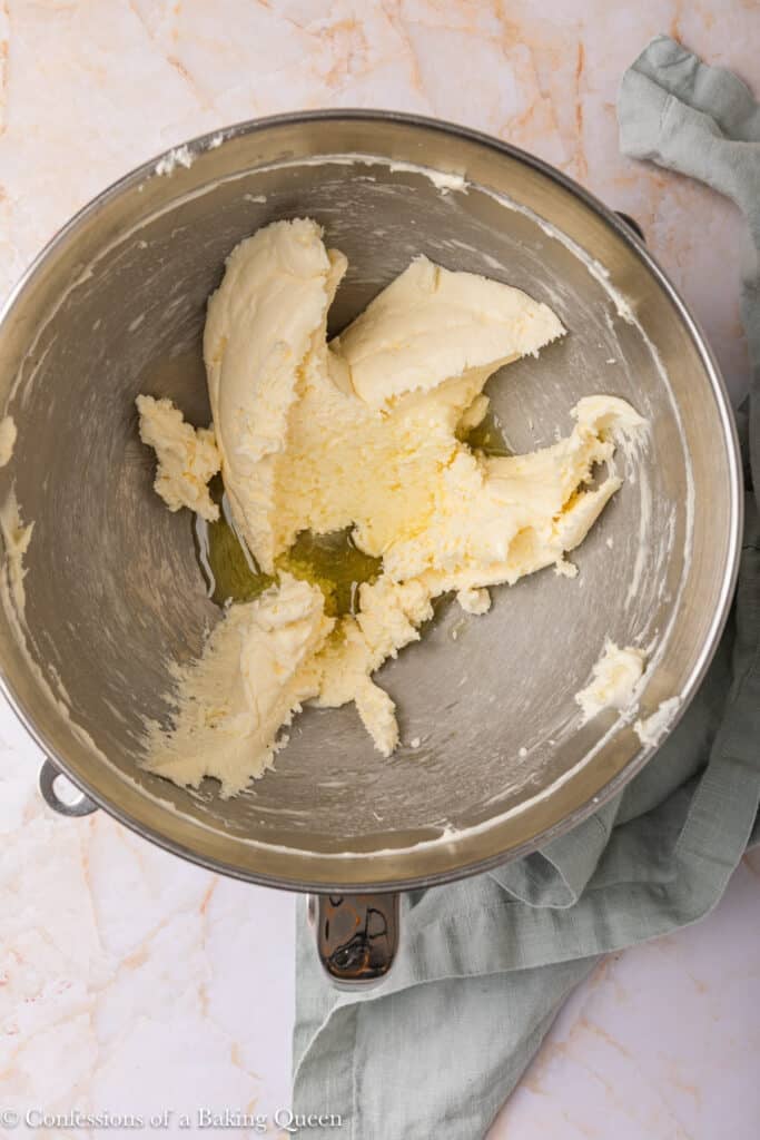 A metal mixing bowl containing partially creamed butter and sugar, with some liquid vanilla extract in the center. The bowl sits on a marble surface next to a folded cloth.