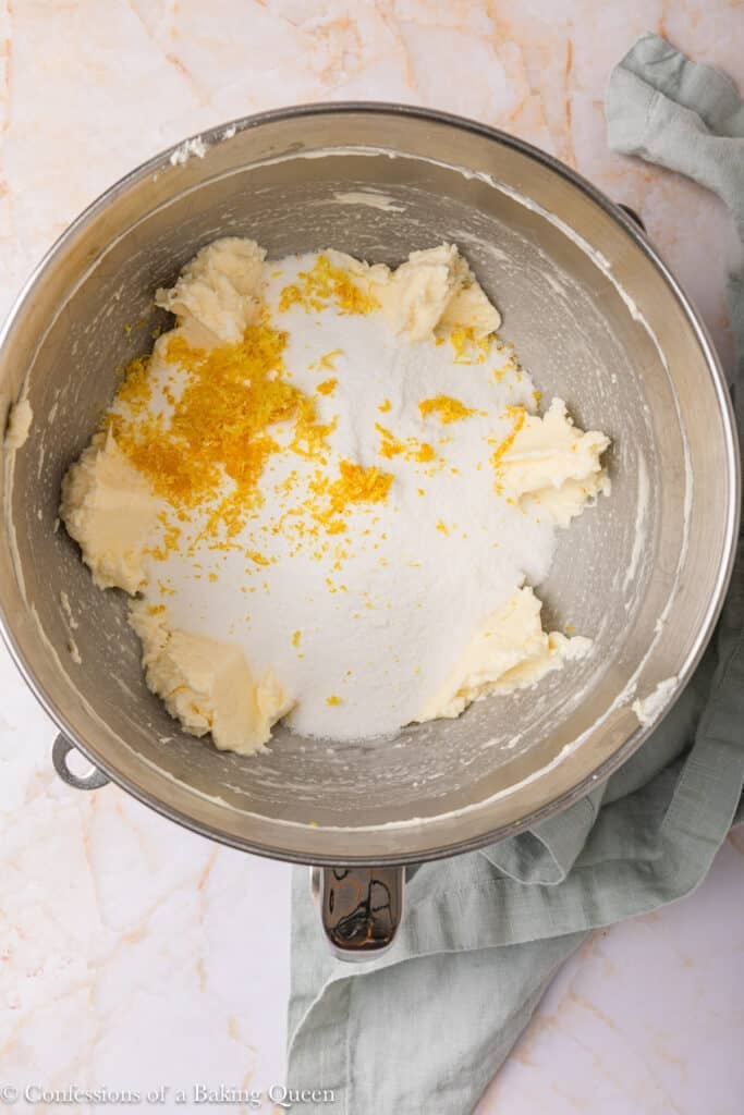 A metal mixing bowl containing butter, granulated sugar, and orange zest on a marble surface with a green cloth beside it.