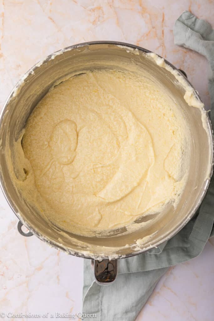 A metal mixing bowl containing creamed butter and sugar mixture sits on a light marble countertop with a green cloth partially visible underneath.