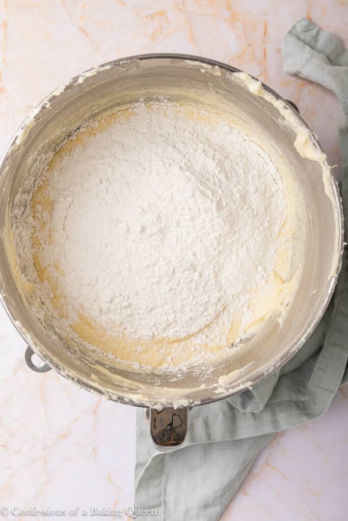 A metal mixing bowl containing flour on top of a creamy batter, set on a marble surface with a light green cloth partially underneath.