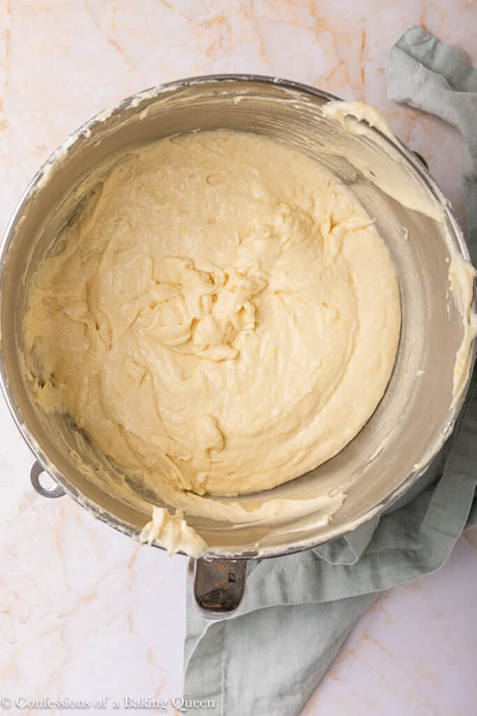 A metal mixing bowl filled with smooth, creamy cake batter sits on a marble surface, partially wrapped with a light green cloth.